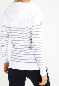 Hoodie blanc avec des rayures horizontales grises, poignets et ourlet côtelés, présentant un petit logo sur la manche et une capuche avec cordon.