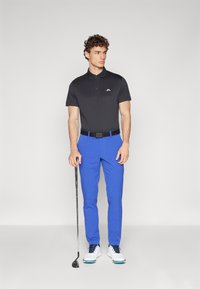Čierne polo tričko s bielym logom, modré nohavice, čierny opasok, drží golfovú palicu, obuté biele a námornícke golfové topánky. Hladká, jemná látka.