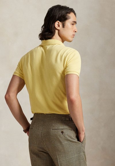 Vyras su tamsiais vidutinio ilgio plaukais dėvi geltoną trumpomis rankovėmis polo marškinėlį ir rudas languotas kelnes, stovintis su rankomis kišenių viduje.