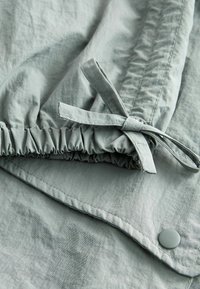 Lichtgrijze stof met een zachte textuur, voorzien van een geplooide elastische band en een strikdetail. Inclusief een gladde knoopaccent.