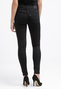 Mujer vistiendo jeans ajustados negros y zapatos de tacón alto negros, mostrada desde atrás sobre un fondo blanco liso.