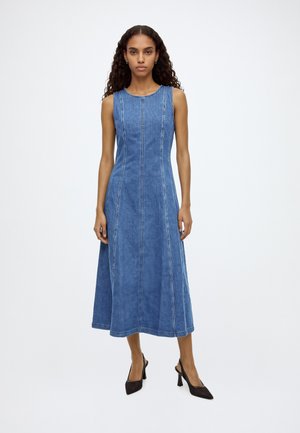 Woman with long curly hair wearing a sleeveless blue denim midi dress and black high-heeled slingback shoes standing against a white background.