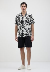 Chemise à manches courtes avec un motif abstrait noir et blanc, associée à un short noir roulé et des baskets blanches, sur un fond neutre.