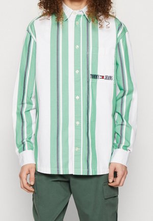Persoon draagt een overhemd met lange mouwen en verticale strepen in groen, wit en grijs, met het "Tommy Jeans" logo op het borstzakje.