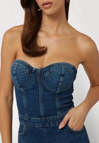 Modrá džínsová top bez ramienok s priliehavým telom, tvarovanými poháromi a prednými švami. Obsahuje vrecko a textúru látky strednej hmotnosti.