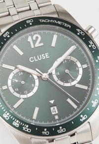 Ατσάλινο ρολόι χειρός με πράσινο καντράν, λευκούς δείκτες ωρών, δύο υποκαντράν, παράθυρο ημερομηνίας στις 6, και κλίμακα ταχύμετρου στη στεφάνη.