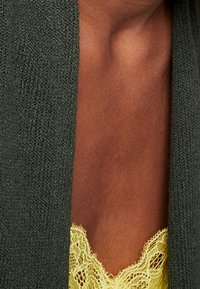 Dunkelgrüne Strickjacke über einer gelben Spitzen-Ärmellose, die die Textur des Stoffs und die filigranen Spitzenverzierungen zeigt.