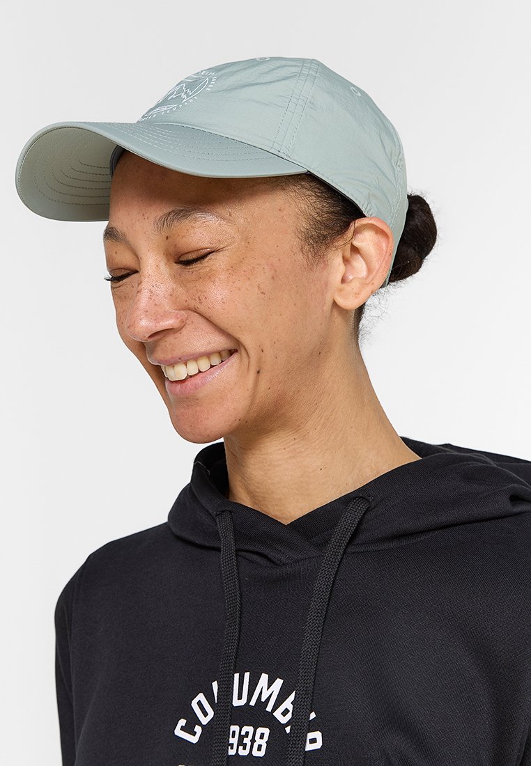 Lächelnde Person mit einer hellgrauen Baseballmütze und einem schwarzen Columbia-Hoodie mit einem Frontlogo, die nach unten schaut und die Augen geschlossen hat.