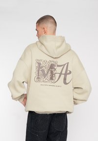 Haut à capuche beige surdimensionné avec un grand motif décoratif "MA" au dos et le texte "MULTIPLY APPAREL CLUB" ; matière douce et design à capuche.