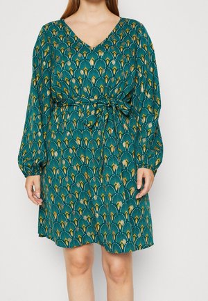 Femme portant une robe à manches longues bleu canard avec un motif en forme d’éventail jaune et blanc et une ceinture à la taille, debout devant un fond uni.