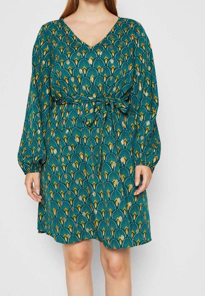 Femme portant une robe à manches longues bleu canard avec un motif en forme d’éventail jaune et blanc et une ceinture à la taille, debout devant un fond uni.