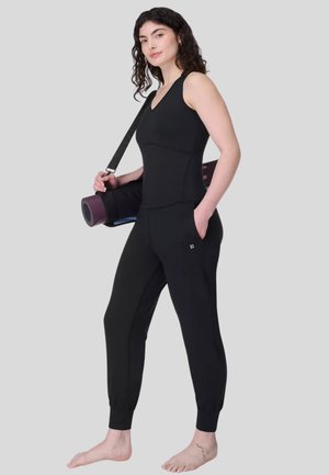 Vrouw in een zwart tanktop en joggingbroek draagt een opgerolde yogamat over haar schouder, staat blootsvoets tegen een effen achtergrond.