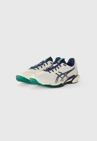 White athletic shoes with navy blue accents, green soles, textured mesh upper, and side logo detailing. Designed for performance and comfort.