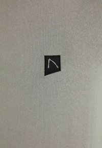 Chemise grise en tricot avec un petit patch noir orné d'un dessin géométrique blanc, présentant une texture lisse et une coupe ajustée.