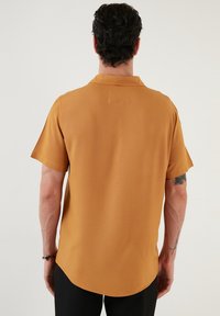 Koszula z krótkim rękawem i kołnierzykiem, wykonana z ciepłej musztardowej tkaniny. Gładka faktura, zaokrąglony dół i luźny krój. Bez widocznych wzorów i elementów metalowych.