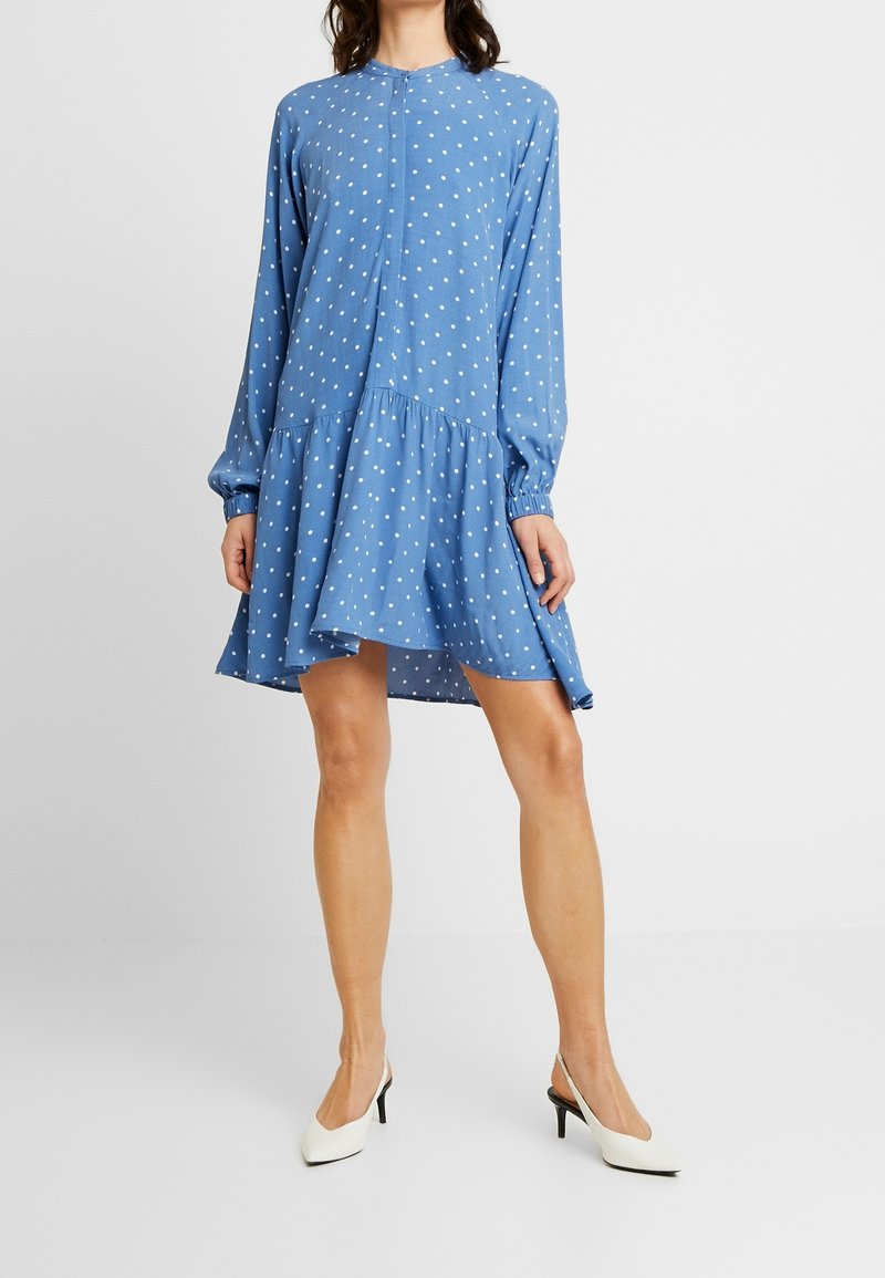 Femme portant une robe bleue à manches longues à pois et des chaussures blanches à talons hauts, debout devant un fond clair uni.