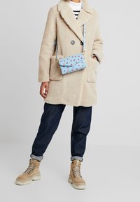 Abrigo de piel sintética beige con grandes bolsillos, llevado sobre una camiseta a rayas blancas y negras, combinado con vaqueros oscuros y botas beige con cordones. Bolso azul con patrón de corazones rojos.