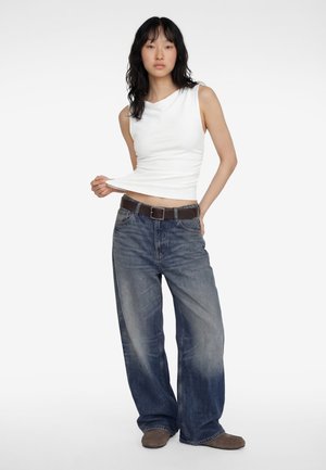 Femme aux longs cheveux noirs portant un débardeur blanc, un jean bleu ample avec une ceinture, et des chaussures marron, debout et tirant sur son haut.