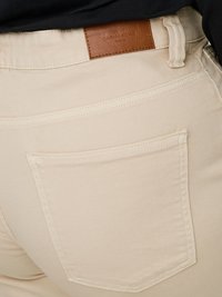 Gros plan de la poche arrière d’un jean beige en denim avec des détails cousus et une étiquette de marque en cuir marron sur la ceinture.