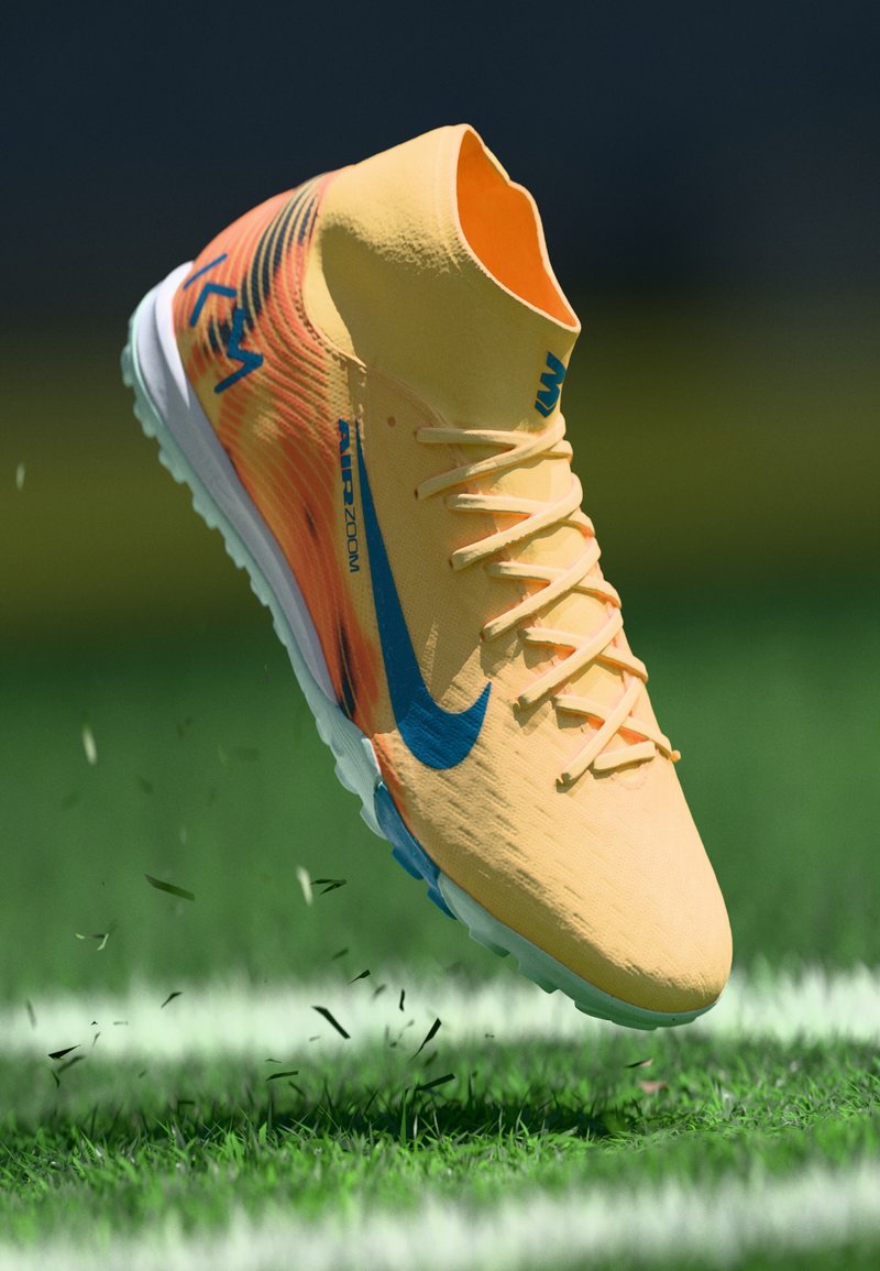 Žluté a oranžové vysoké fotbalové kopačky s modrým Nike swooshem ve vzduchu nad zelenou trávou s bílou čárou na hřišti, ukazující pohyb a částice trávy.