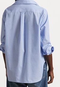 Persona che indossa una camicia a maniche lunghe azzurra con le maniche arrotolate e jeans blu, vista da dietro su uno sfondo bianco.