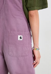 Salopette in cotone lavanda con una tasca rettangolare che presenta il logo Carhartt. Il tessuto è liscio e ha una finitura opaca.