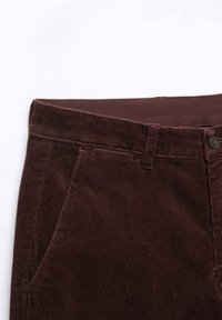 Gros plan de la ceinture d’un pantalon en velours côtelé marron avec poche latérale, bouton et passants visibles sur fond blanc.