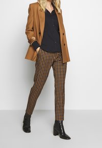 Bruine blazer met zwarte knopen, zwart overhemd met knopen en bruine geruite broek. Zwarte enkellaarsjes met hak en een gestructureerde afwerking.