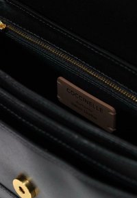 Interno di una borsa in pelle nera con una cerniera dorata, che mostra un'etichetta con la scritta "Coccinelle - Vera Pelle Italiana." Texture liscia, design minimale.