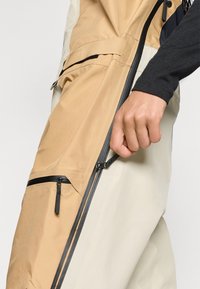 Beige és krémszínű külső viselet, fél cipzáros kialakítással, több cipzáras zsebbel és textúrált felülettel. Egy kéz a cipzár állításával foglalkozik.