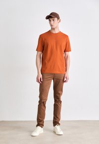 Brun baseball cap, oransje bomull t-skjorte med lite logo, lys beige tettsittende bukser og hvite sneakers. Står på en nøytral gulv.