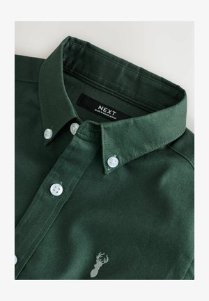 Camisa de algodón verde con cuello puntiagudo, botones blancos y un pequeño logo de ciervo bordado en gris claro en el frente. Etiqueta visible en el interior.