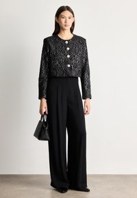 Giacca nera corta con motivo in pizzo argento, caratterizzata da tre grandi bottoni, abbinata a pantaloni a gamba larga neri a vita alta e una piccola borsa.