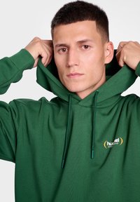 Grön hoodie gjord av mjukt tyg med en dragsko i huvan. Har en liten logotyp i gult och vitt på bröstet. Enkel och avslappnad design.