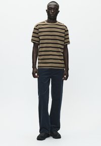 Brun og beige stribet t-shirt med korte ærmer, parret med mørkeblå lige ben jeans og sorte sko, sat mod en ensfarvet hvid baggrund.
