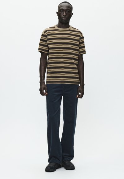 T-shirt a strisce marroni e beige con maniche corte, abbinato a jeans straight leg blu scuro e scarpe nere, su sfondo bianco semplice.