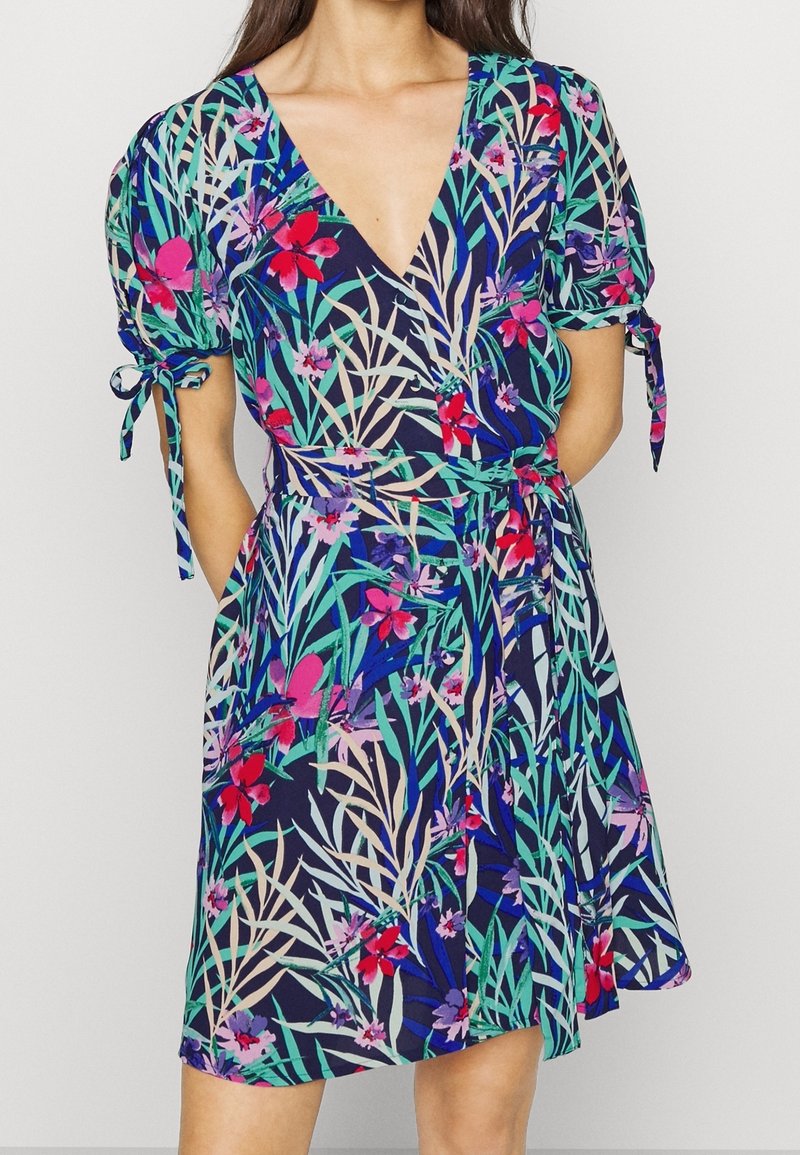 Femme portant une robe portefeuille mi-longue aux motifs floraux tropicaux vibrants en bleu, vert, rouge et violet avec de courtes manches bouffantes nouées sur les côtés.
