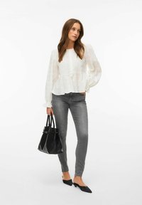 Femme portant un chemisier blanc à manches longues et un jean skinny gris, tenant un sac à main noir, debout avec une main dans la poche sur fond blanc.