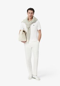 Bílá polokošile s logem Lacoste, krémová pletená šála, krémová taška, bílé kalhoty na míru a bílé tenisky se zelenými akcenty.