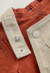 Koszula w kolorze rdzy z czarnym nadrukiem palm, zestawiona z beżowymi ogrodniczkami z tkaniny, zapinanymi na metalowe zatrzaski i ozdobionymi okrągłą metką z logo.