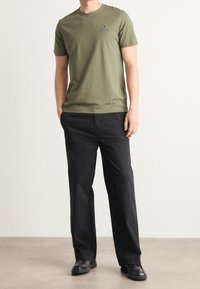Camiseta de algodón verde oliva con cuello redondo y mangas cortas, con un pequeño logo. Combinada con pantalones negros y zapatos oscuros.