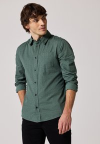 Zelená košile s vzorem, s límečkem, černými knoflíky a dlouhými rukávy, s kapsou na levé straně hrudi a vyhrnutými manžetami.
