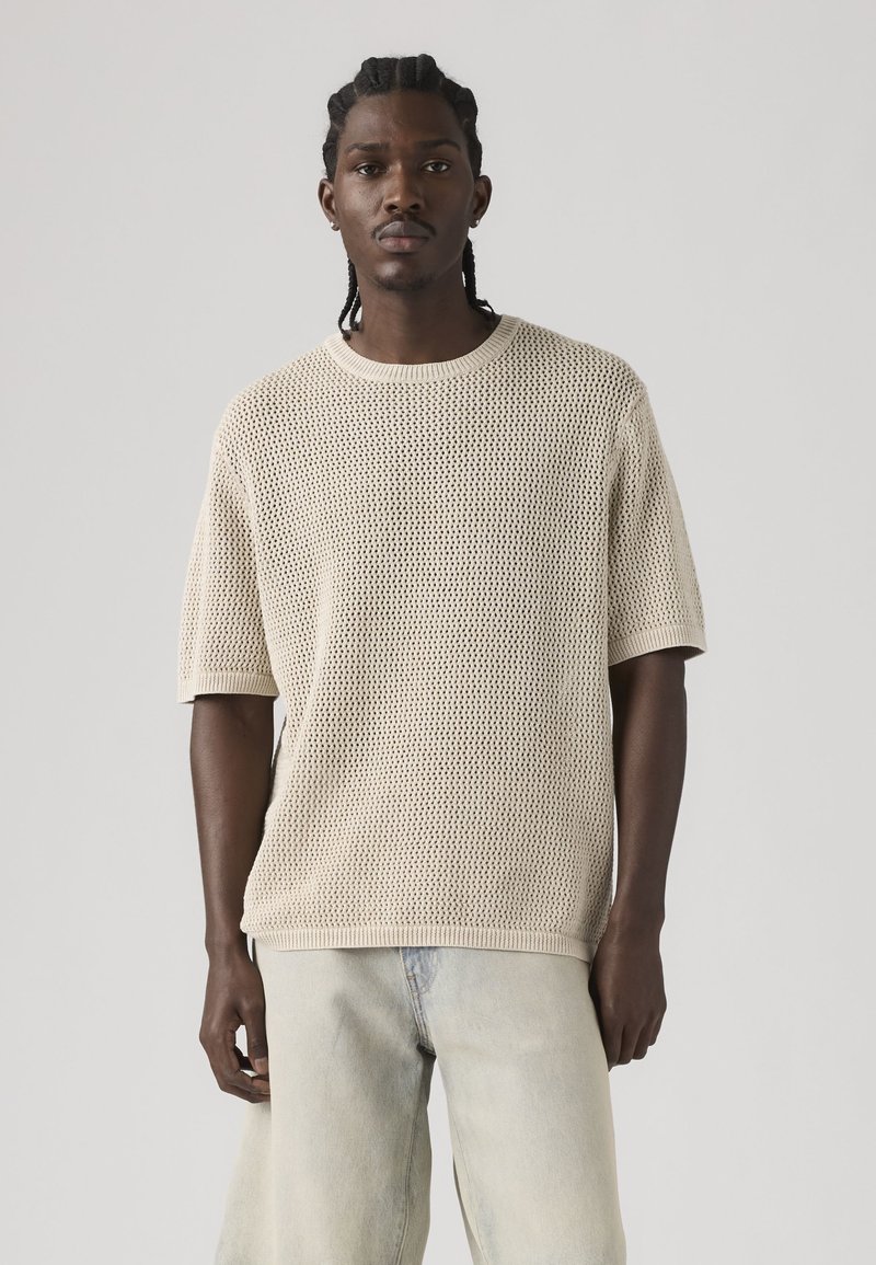 Jeune homme aux cheveux tressés portant un pull beige en tricot à manches courtes et un jean bleu clair sur un fond gris uni.