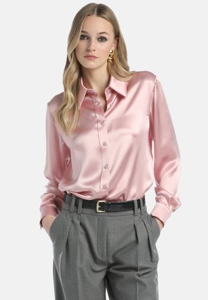 Camicia rosa in satin con bottoni e colletto, caratterizzata da maniche lunghe. Abbinata a pantaloni grigi plissettati e una cintura nera. Texture liscia e lucida.