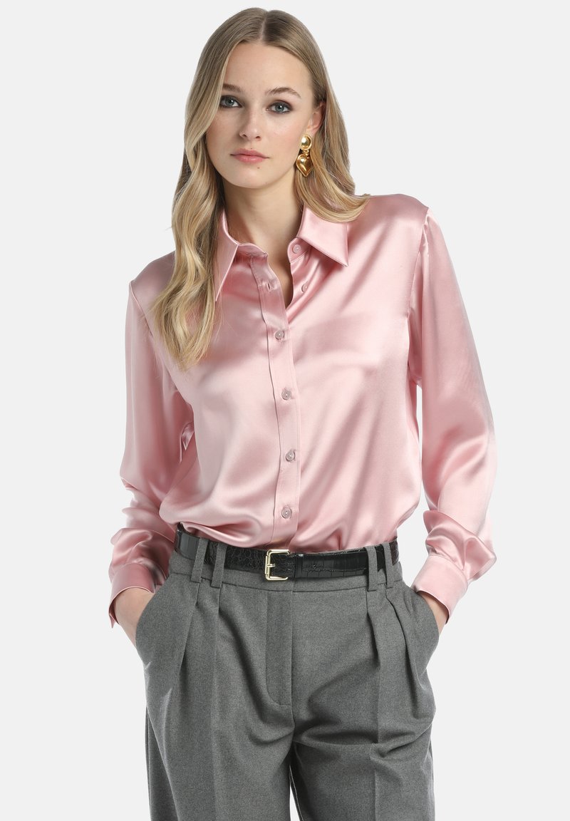 Rosa satinierte Hemdbluse mit Kragen und langen Ärmeln. Kombiniert mit grauen plissierten Hosen und einem schwarzen Gürtel. Glatte, glänzende Textur.
