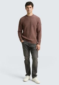 Bruine grofgebreide trui met ronde hals, gecombineerd met grijze spijkerbroek en witte sneakers; staat tegen een neutrale achtergrond, handen ontspannen langs het lichaam.