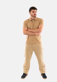 Polo shirt color sabbia con colletto, pantaloni cargo beige con tasche e sneakers nere con dettagli color sabbia. Il modello sta in piedi con le braccia incrociate su uno sfondo bianco.