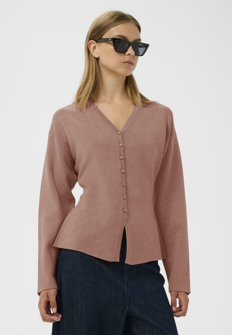 Rosenfarbener Knopf-Cardigan mit langen Ärmeln, V-Ausschnitt, strukturiertem Stoff und seitlichen Taschen. Über dunklen Denim-Hosen getragen.