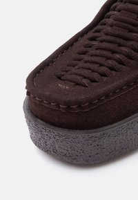 Gros plan sur la pointe d'une chaussure en daim marron foncé avec un détail supérieur tissé et une semelle texturée noire sur fond blanc.