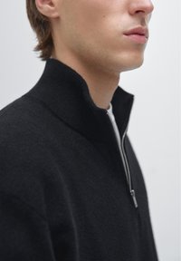 Zwarte zip-up trui gemaakt van zachte stof met een hoge kraag, voorzien van een zilveren rits, een gladde textuur en een getailleerd ontwerp.